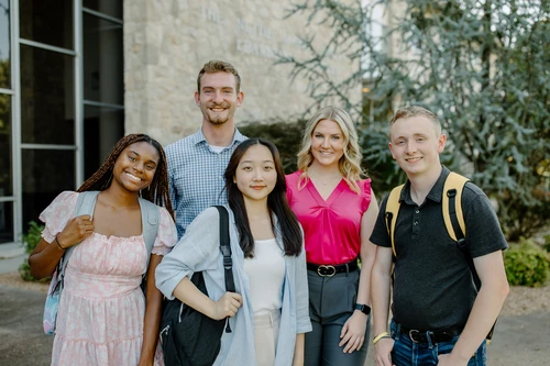 College students pose with backpacks in front of academic building. 